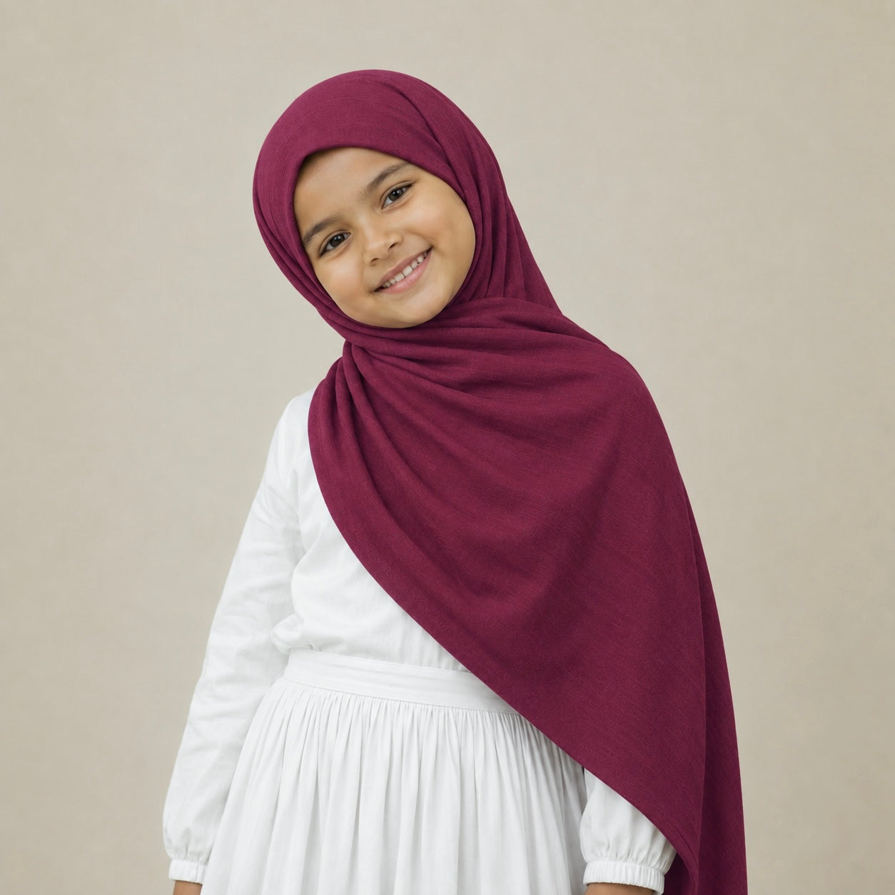 A young girl wearing a deep burgundy Kids Jersey hijab and a white long-sleeve dress, posing against a solid light beige background.